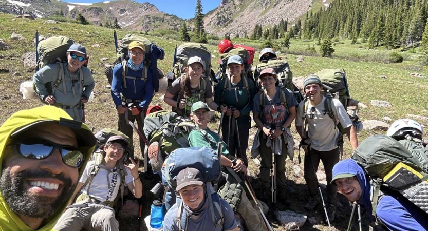 Instructors and students wearing backpacks pose for a group photo in a green alpine meadow 